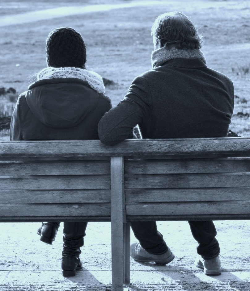 Two people sit side by side on a wooden park bench, facing away, wearing winter coats and scarves.